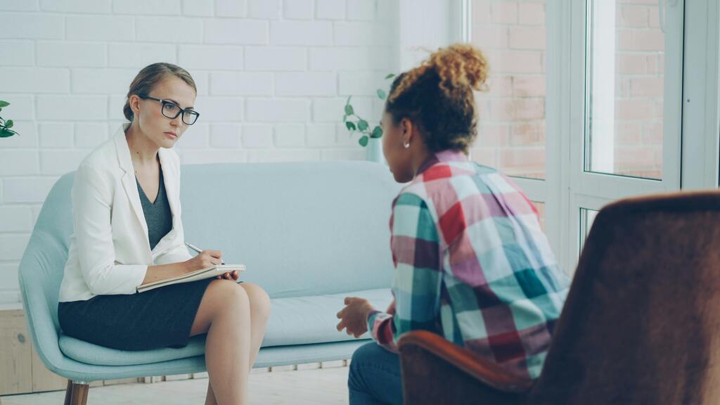 Female therapist with glasses in a white blazer sits on a blue couch, taking notes in a counseling session with a client wearing a colorful plaid shirt across from her in a bright room.